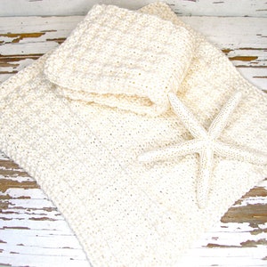 May include: Cream-colored, crocheted washcloths and a larger square blanket are displayed with a white starfish. The items are arranged on a distressed, white-painted wooden surface, creating a beachy, spa-like aesthetic. The crochet work has a textured appearance.