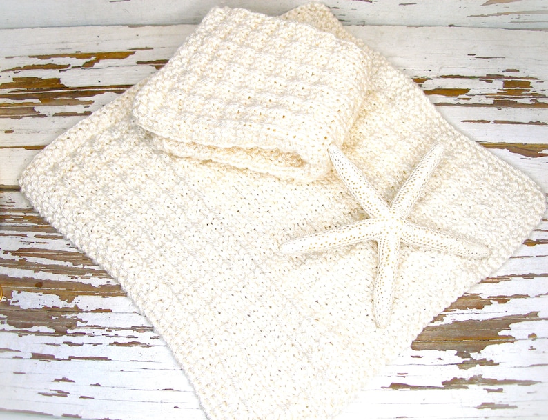 May include: Cream-colored, crocheted washcloths and a larger square blanket are displayed with a white starfish. The items are arranged on a distressed, white-painted wooden surface, creating a beachy, spa-like aesthetic. The crochet work has a textured appearance.