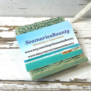 May include: A turquoise ceramic soap dish with a textured surface, holding a business card. The card reads "SeamariesBounty" and lists website addresses. A white starfish rests in the background on a distressed white wooden surface.