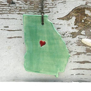 May include: A green ceramic pendant in the shape of the state of Georgia, with a red heart in the center. The pendant hangs from a dark chain. A white starfish rests on a weathered wooden surface.
