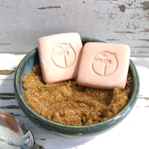 May include: Two square, peach-colored soaps with the word "smile" and a peach design are nestled in a small, teal-rimmed bowl filled with brown sugar. The bowl sits on a white, weathered wooden surface.