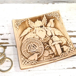 May include: A square beige tile featuring a detailed relief carving of a bearded figure wearing a winged helmet and an eye patch, holding a sword and a shield with a triangular symbol, flanked by two birds, alongside three gold rings and red earrings.