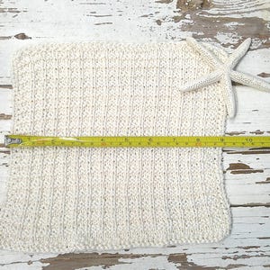 May include: Cream-colored knitted square with a textured pattern, measuring approximately 14 inches across, with a white starfish and a yellow measuring tape. The item is displayed on a distressed white wooden surface.