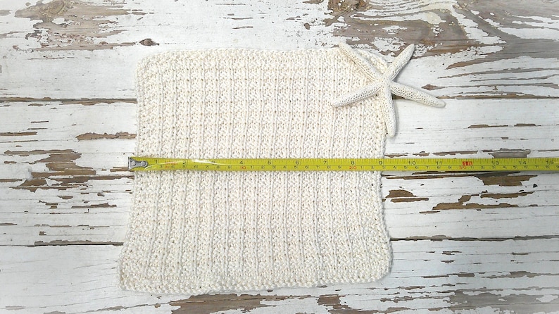 May include: Cream-colored knitted square with a textured pattern, measuring approximately 14 inches across, with a white starfish and a yellow measuring tape. The item is displayed on a distressed white wooden surface.