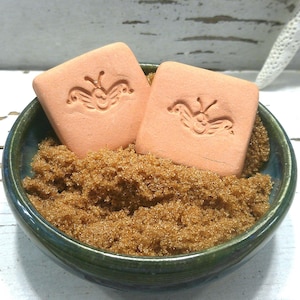 May include: Two square, peach-colored clay pieces, each embossed with a butterfly design, are nestled into a small, dark green ceramic bowl filled with coarse, golden-brown granules. The bowl has a smooth, rounded rim and a deep, dark interior.
