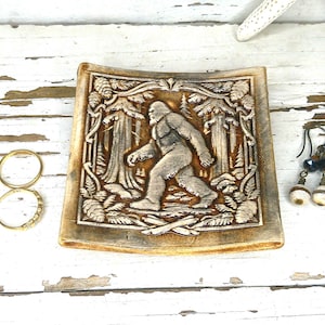 May include: A square ceramic trinket dish featuring a relief carving of a Bigfoot creature walking through a forest, accompanied by three gold-toned metal rings and a pair of dangling earrings with dark beads and white spherical accents.