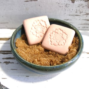 May include: A small, round, dark green ceramic bowl filled with brown sugar. Two square, light peach-colored soap bars, each embossed with a detailed floral design, are resting on top of the brown sugar within the bowl.