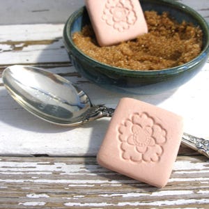 May include: A silver spoon and a small teal bowl filled with brown sugar sit on a distressed white wooden surface. Two square, light pink soaps with a floral design are also present, one in the bowl and one next to the spoon.
