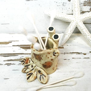 May include: A whimsical ceramic container shaped like a crab, holding cotton swabs. The container is tan with brown spots and black eyes. A white starfish and cotton swabs are also in the image.