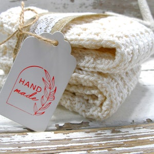 May include: A stack of off-white, crocheted washcloths tied with twine and a tag. The tag reads "HAND made" in red script with a leaf design. A white starfish is in the background.