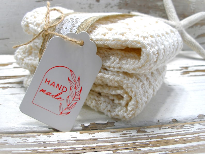 May include: A stack of off-white, crocheted washcloths tied with twine and a tag. The tag reads "HAND made" in red script with a leaf design. A white starfish is in the background.