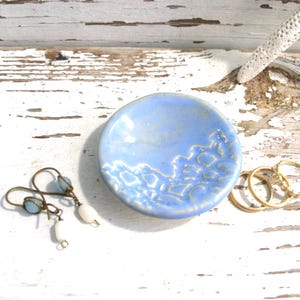 Peut inclure: Un petit plat &agrave; bijoux en c&eacute;ramique bleu clair avec un motif en forme de dentelle. Il contient une paire de boucles d'oreilles avec des perles bleues et des pendentifs blancs en forme de goutte, et trois bagues dor&eacute;es. Sur une surface en bois blanc vieilli.