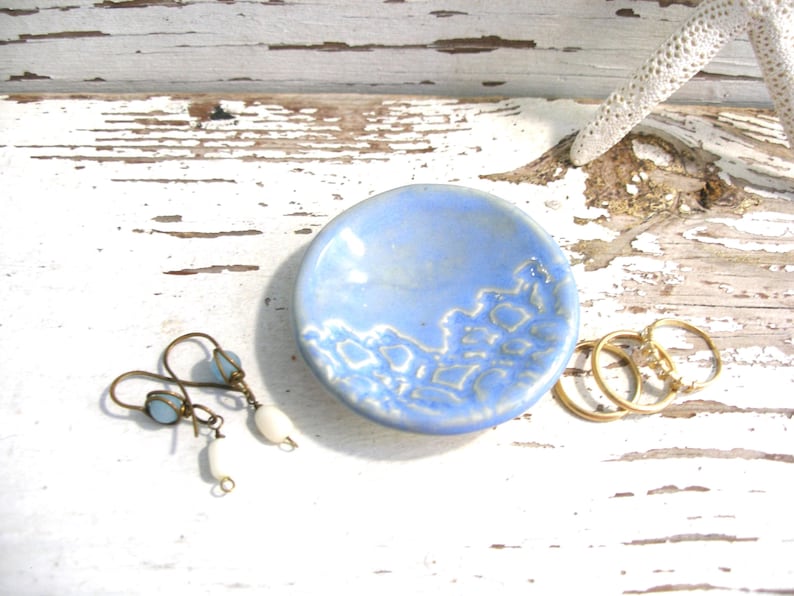 Peut inclure: Un petit plat &agrave; bijoux en c&eacute;ramique bleu clair avec un motif en forme de dentelle. Il contient une paire de boucles d'oreilles avec des perles bleues et des pendentifs blancs en forme de goutte, et trois bagues dor&eacute;es. Sur une surface en bois blanc vieilli.
