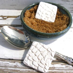 May include: A dark teal ceramic bowl filled with brown sugar and a white square textured sugar saver, accompanied by a silver spoon featuring an ornate handle and a matching white square textured piece attached to the spoon neck.