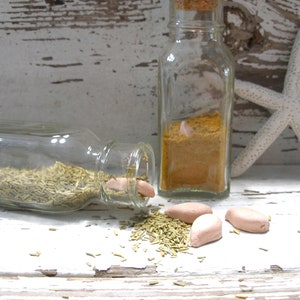 May include: A still life arrangement featuring clear glass bottles filled with spices and herbs. One bottle is tipped over, spilling dried rosemary. Three small, light-colored, oblong objects are scattered nearby. A starfish rests in the background.