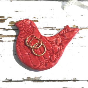 May include: A red ceramic bird-shaped ring holder with a textured surface. Three gold rings are placed on the holder. The ring holder is on a white, distressed wooden surface.