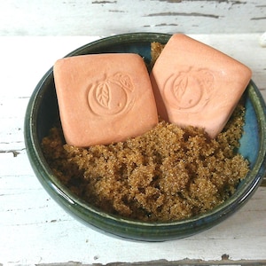 May include: A small, round, dark teal bowl filled with brown granules and two square, peach-colored soaps. Each soap has a carved peach design. The bowl sits on a white, distressed wooden surface.