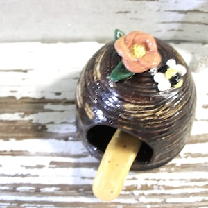 May include: A ceramic beehive-shaped incense holder. The brown holder has a textured surface and a small opening for the incense stick. It is decorated with a peach-colored flower, green leaves, and a small bee.