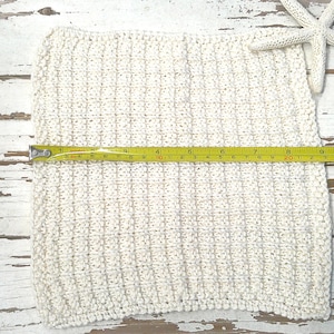 May include: Cream-colored, square, knitted washcloth with a yellow measuring tape showing a length of approximately 11 inches. A white starfish rests on the upper right corner. The washcloth is on a distressed white wooden surface.