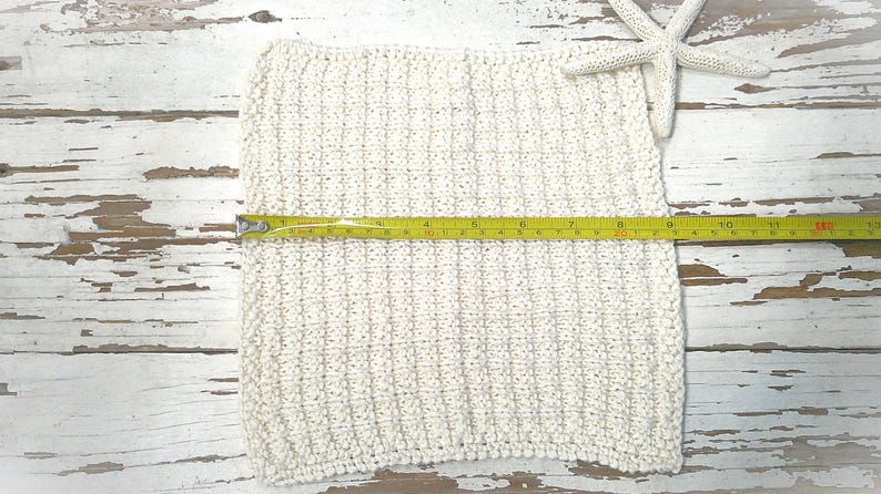 May include: Cream-colored, square, knitted washcloth with a yellow measuring tape showing a length of approximately 11 inches. A white starfish rests on the upper right corner. The washcloth is on a distressed white wooden surface.