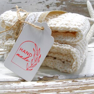 May include: A stack of off-white, crocheted washcloths tied with twine and lace, with a tag that reads "HAND made" in red. A starfish rests nearby on a distressed, white wooden surface. The washcloths are folded neatly.
