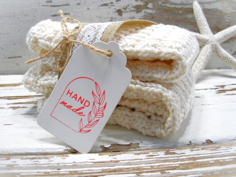 May include: A stack of off-white, crocheted washcloths tied with twine and lace, with a tag that reads "HAND made" in red. A starfish rests nearby on a distressed, white wooden surface. The washcloths are folded neatly.
