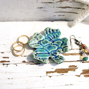 May include: A small, irregular, clover-shaped ceramic dish with a glossy, mottled blue and green glaze. To the left are three thin, plain gold-toned metal rings. To the right are two dangling earrings with bronze-toned hooks and beads.