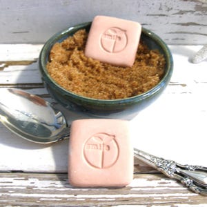 May include: A dark green ceramic bowl filled with brown granulated sugar, accompanied by a silver spoon and two square, light pink soap bars. Each soap bar features a circular embossed logo with the word smile and a small leaf icon on top.
