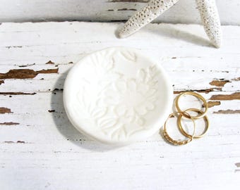May include: A small, round, white ceramic dish with a floral design, holding three gold rings. The dish is on a distressed white wooden surface, with a white starfish in the background.