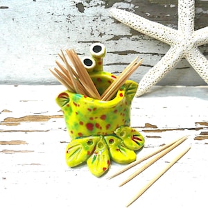 May include: A whimsical, hand-painted ceramic crab toothpick holder in shades of yellow and green with red spots. The crab has large, googly eyes and holds wooden toothpicks. A white starfish rests nearby, adding a coastal theme.
