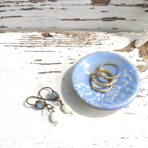 Peut inclure: Un petit plat rond en c&eacute;ramique bleu clair avec un motif textur&eacute; contient trois bagues dor&eacute;es. Une paire de boucles d'oreilles avec des perles bleues et blanches repose &agrave; proximit&eacute;. Les articles sont pr&eacute;sent&eacute;s sur une surface en bois blanc vieilli.