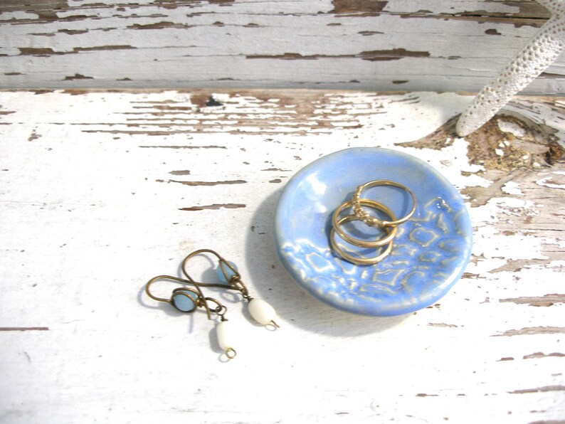 Peut inclure: Un petit plat rond en c&eacute;ramique bleu clair avec un motif textur&eacute; contient trois bagues dor&eacute;es. Une paire de boucles d'oreilles avec des perles bleues et blanches repose &agrave; proximit&eacute;. Les articles sont pr&eacute;sent&eacute;s sur une surface en bois blanc vieilli.