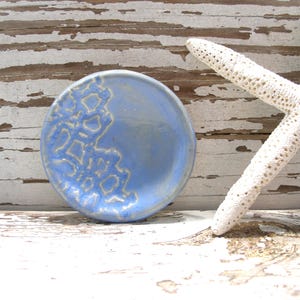Peut inclure: Un petit plat rond en c&eacute;ramique bleu clair avec un motif textur&eacute; blanc cass&eacute; sur un c&ocirc;t&eacute;. Une &eacute;toile de mer blanche repose &agrave; c&ocirc;t&eacute; du plat sur une surface en bois vieilli peint en blanc. Le plat mesure environ 10 cm de diam&egrave;tre.