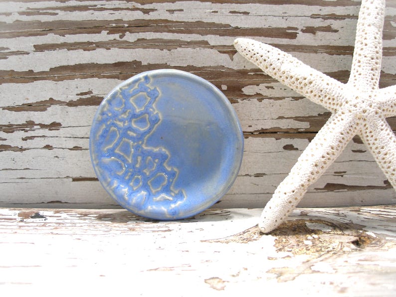 Peut inclure: Un petit plat rond en c&eacute;ramique bleu clair avec un motif textur&eacute; blanc cass&eacute; sur un c&ocirc;t&eacute;. Une &eacute;toile de mer blanche repose &agrave; c&ocirc;t&eacute; du plat sur une surface en bois vieilli peint en blanc. Le plat mesure environ 10 cm de diam&egrave;tre.