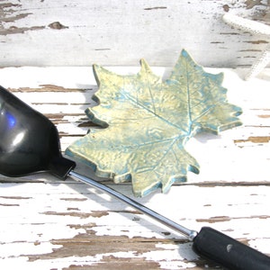 May include: A black plastic kitchen ladle with a long metal handle and a ceramic spoon rest shaped like a maple leaf with a textured, mottled blue and yellow glaze. The spoon rest features visible leaf veins and jagged edges.