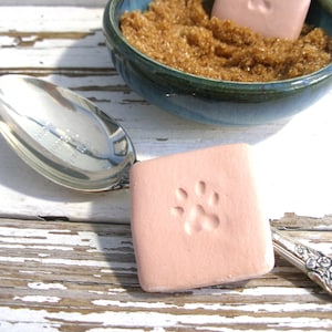 May include: A square, light pink soap with a paw print is next to a silver spoon and a teal bowl filled with brown sugar. The items are arranged on a distressed white wooden surface.