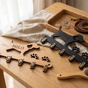 Puede incluir: Dos soportes de pared para correas hechos de madera, uno en acabado marrón claro natural con el nombre MILO y otro en acabado negro con el nombre LUNA. Cada soporte presenta una silueta de perro, cuatro ganchos metálicos y dos huellas.