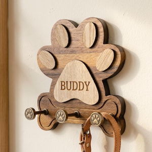 Peut inclure: Un porte-laisse mural en bois en forme de patte avec une base en forme d'os. Le bois foncé présente des accents en bois clair sur les coussinets, avec le nom BUDDY gravé sur le coussinet central. Trois crochets en bronze avec des détails de patte tiennent une laisse en cuir beige.