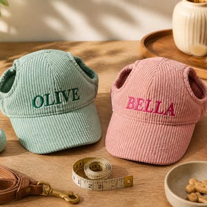Puede incluir: Dos gorras de pana para mascotas con orificios para las orejas, una verde claro con el nombre bordado OLIVE y una rosa empolvado con el nombre bordado BELLA. Una cinta métrica blanca marca 150 cm, junto a una correa marrón y premios.