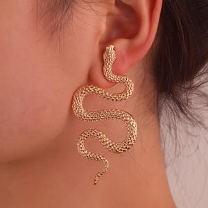 Peut inclure: Une boucle d'oreille en métal doré en forme de serpent sinueux avec une surface texturée rappelant des écailles. Le bijou présente un corps courbé qui pend sous le lobe de l'oreille, avec la tête du serpent située en haut près du perçage.