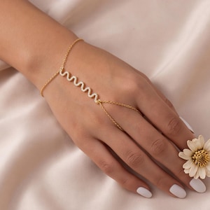 Puede incluir: Una pulsera de mano dorada que presenta una delicada barra ondulada incrustada con pequeñas piedras transparentes. La cadena se extiende desde una pulsera hasta un anillo que rodea el dedo medio, con un diseño de eslabones metálicos finos.