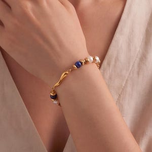 Könnte beinhalten: Ein goldfarbenes Kettenarmband mit abwechselnden weißen Perlen, tiefblauen Lapislazuli-Perlen und kleinen goldenen Zwischenelementen. Das Armband verfügt über ein zentrales, geschwungenes goldenes Element und einen sicheren Verschluss.