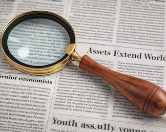 May include: A magnifying glass with a circular gold-toned metal frame and a polished brown wooden handle. The lens is positioned over a newspaper page featuring the visible text "Assets Extend World Wi" and "Senior economists" in black ink.