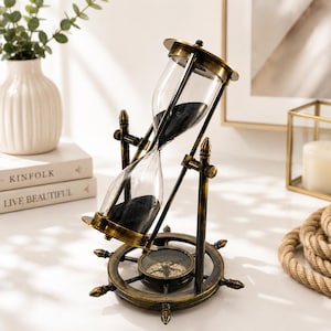 Puede incluir: Un reloj de arena de latón de estilo antiguo con arena negra, montado sobre un marco giratorio sostenido por una base circular con forma de timón de barco. La base incluye una brújula integrada en el centro, con detalles metálicos.