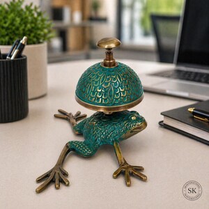 May include: A metal desk bell featuring a frog base with a textured teal and bronze finish. The bell dome has a repeating scale-like pattern and a small knob on top. The frog is depicted in a crouching position with extended legs and webbed feet.