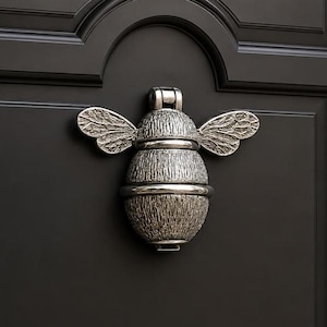 May include: A silver-toned metal door knocker shaped like a bee with textured wings and a segmented body. The bee is mounted on a dark, matte-finished door with a recessed panel design. The metal surface features a weathered, antique-style finish.