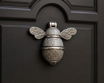 May include: A silver-toned metal door knocker shaped like a bee with textured wings and a segmented body. The bee is mounted on a dark, matte-finished door with a recessed panel design. The metal surface features a weathered, antique-style finish.