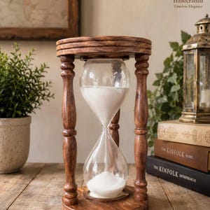 May include: A wooden hourglass featuring a dark brown finish with turned pillars connecting a circular top and base. The clear glass bulb contains white sand, with a portion of the sand currently resting in the bottom chamber of the device.