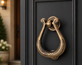 May include: A brass-toned metal door knocker with a lyre-inspired shape and ornate detailing, mounted on a matte black paneled door. The knocker features a circular base plate and a curved, textured handle that hangs vertically against the door surface.
