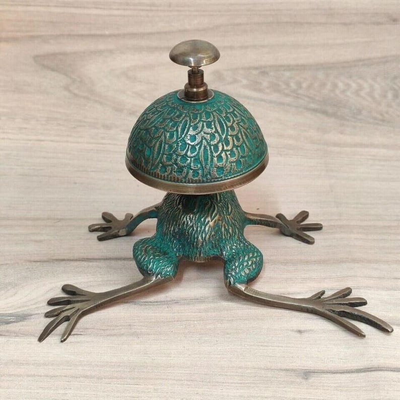 May include: A metal desk bell featuring a frog-shaped base with extended legs and a dome-shaped top. The object has a weathered teal and bronze finish with intricate textured patterns on the dome and the body of the frog, topped with a small knob.